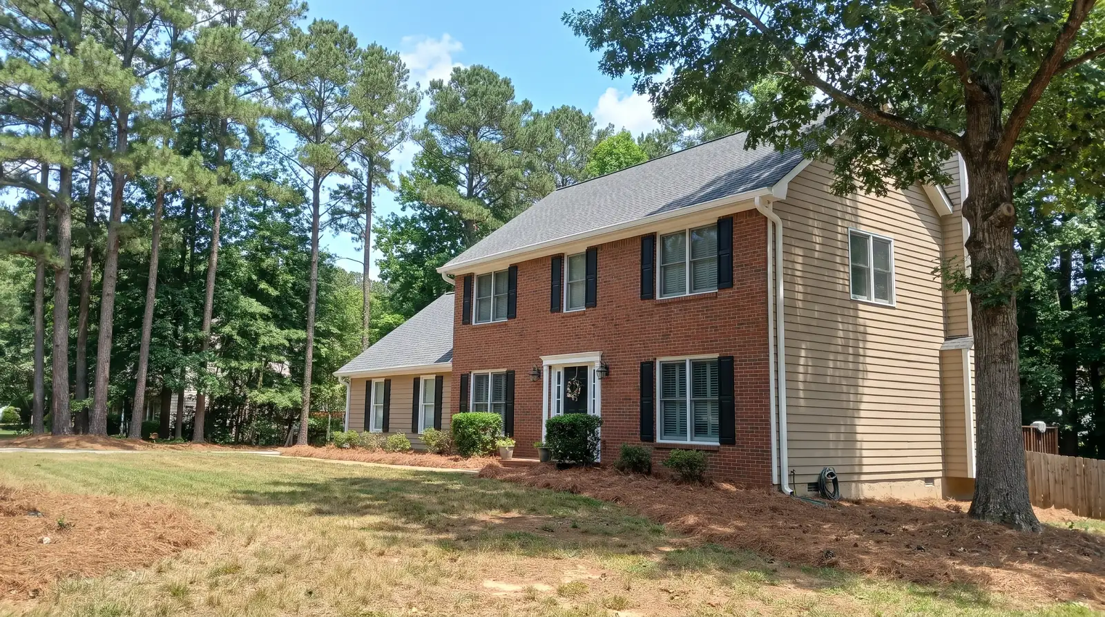 Two-story brick and tan siding house with white gutters and downspouts under tall pines